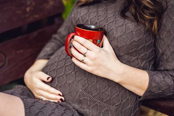 Vientre de una mujer embarazada sentada en un columpio, con una taza de té. mujer embarazada relajándose en el parque . — Foto de Stock