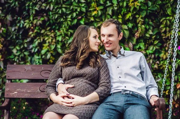 En gravid ung kvinne og mannen hennes. En lykkelig familie som sitter på en huske og holder i magen. gravid kvinne som slapper av i parken . – stockfoto