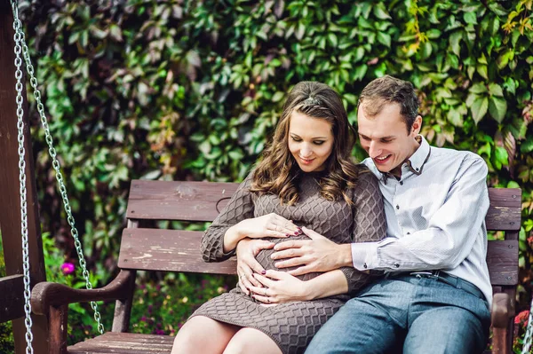 En gravid ung kvinne og mannen hennes. En lykkelig familie som sitter på en huske og holder i magen. gravid kvinne som slapper av i parken . – stockfoto