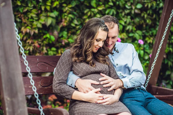 A pregnant young woman and her husband. A happy family sitting on a swing, holding belly. pregnant woman relaxing in the park. — Stock Photo, Image