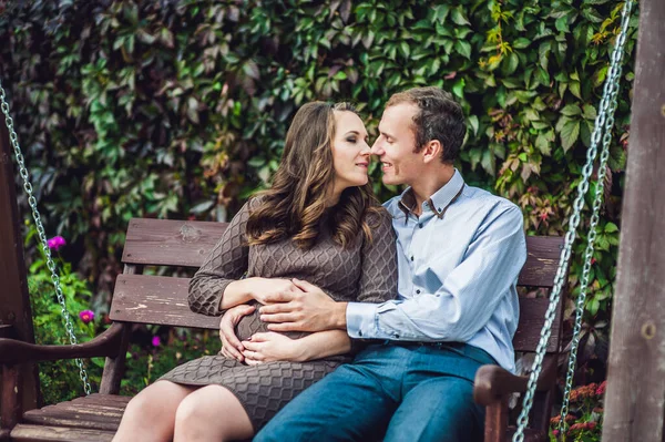 妊娠中の若い女性とその夫。幸せな家族のスイングに座って、腹を保持します。妊娠中の女性は公園でリラックスして. — ストック写真