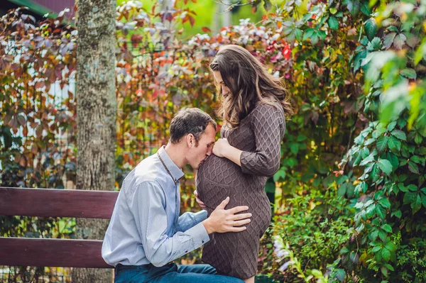 Беременная молодая женщина с мужем. Счастливая семья, стоящая у красной осенней изгороди, будущий отец, слушая это в животе. беременная женщина отдыхает в парке . — стоковое фото