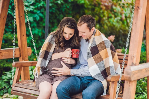 Těhotná mladá žena a její manžel. Šťastná rodina sedící na houpačce a držící se za břicho. těhotná žena relaxace v parku. — Stock fotografie