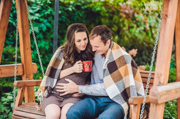 Μια έγκυος νεαρή γυναίκα και ο σύζυγός της. Μια ευτυχισμένη οικογένεια που κάθεται σε μια κούνια, κρατώντας την κοιλιά. έγκυος γυναίκα χαλαρώνοντας στο πάρκο. — Φωτογραφία Αρχείου