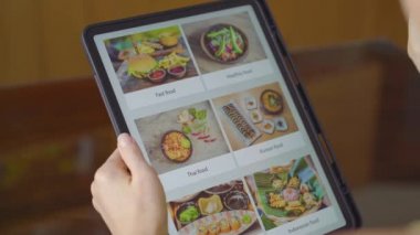 Genç bir kadın, tablet kullanarak internetten yemek sipariş ediyor. Dairesinde bir mutfakta oturuyor.