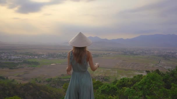 Genç bir kadın turist, vadi manzaralı bir dağın tepesinde duruyor. Asya şapkası takıyor. — Stok video