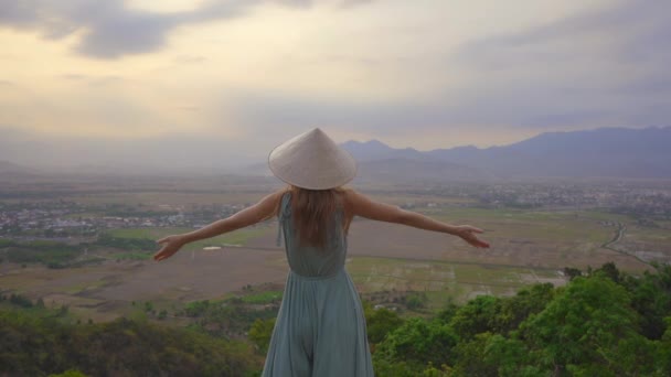 Una giovane turista si trova su una cima di montagna con una vista mozzafiato su una valle. Lei indossa un cappello asiatico — Video Stock