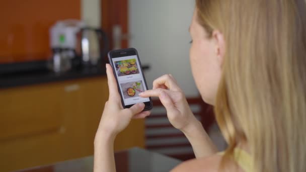 Jonge vrouw bestelt online eten met een smartphone. Ze zit in haar keuken in haar appartement. — Stockvideo