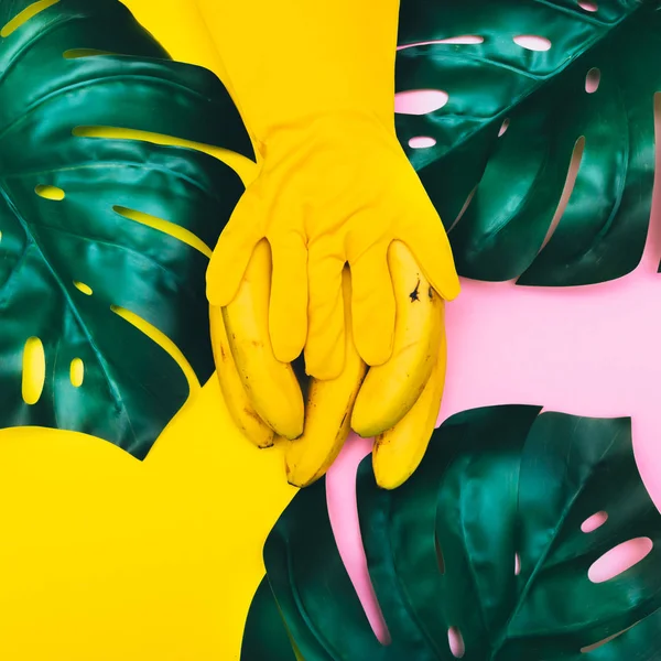 hand in yellow glove is holding  bananas with palm tropical leaves. minimal flat lay. pastel colors. fashion exotic fruit