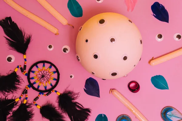 Fortune telling ball with many eyes, a dream catcher, glittering candles, a ring and feathers on coral pink background. Flat lay, modern abstract pop art.