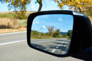 Autumn road in the car mirror. Sunny autumn day and suburban highway. Colorful leaves of trees along the autumn road, colors of leaf-fall. Autumnal landscape. clipart