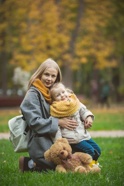 Madre e hija pequeña rubia con el oso de peluche sentado en la hierba verde en el parque de otoño, de cerca — Foto de Stock