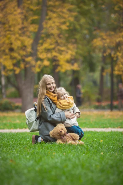 Madre e figlia piccola bionda con orsacchiotto posti a sedere su erba verde nel parco autunnale — Foto Stock