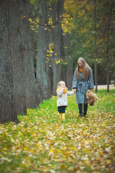 Madre e hija pequeña rubia con oso de peluche caminando en el parque de otoño, vidas amarillas alrededor — Foto de Stock