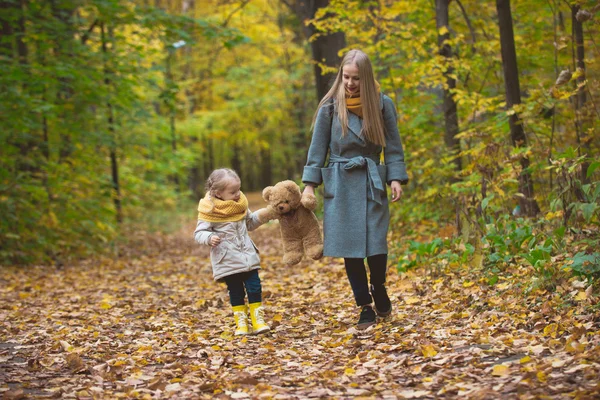 Happy family walking in autumn park - The mother, her daughter and toy Teddy bear among the yellow leaves