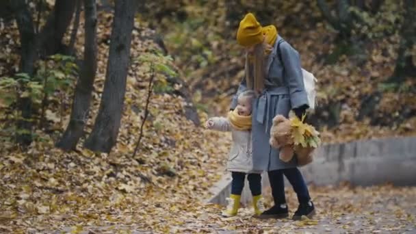 Una linda niña con el pelo rubio y su madre caminando en el parque de otoño y viendo algo, hojas amarillas alrededor — Vídeos de Stock