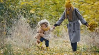 Anne ve kızının oynarken küçük kız sonbahar bir parkta - anne bir sulak tutarak ve sarı yapraklar tekme çocuk verir.