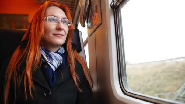 Aantrekkelijke jonge vrouw met rood haar en een bril kijken uit het raam van een trein — Stockvideo