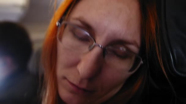 Молодая женщина с рыжими волосами и очками спит в летающем самолете, затем просыпается и улыбается — стоковое видео