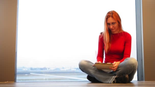 Jovem mulher atraente com cabelo vermelho e óculos usam gadget perto da janela no aeroporto — Vídeo de Stock