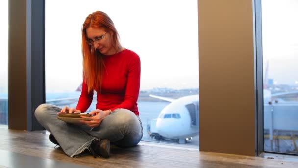 Junge attraktive Frau mit roten Haaren und Brille benutzt Gadget am Fenster des Flughafens vor Flugzeugen — Stockvideo