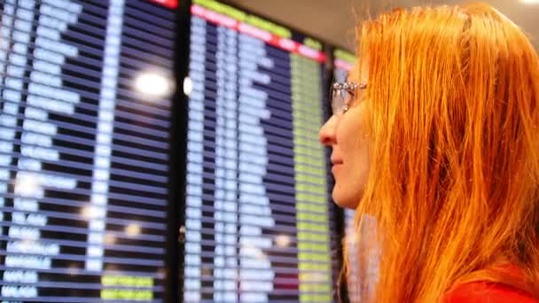 Молодая европейская женщина с рыжими волосами смотрит на расписание вылета в аэропорту — стоковое видео