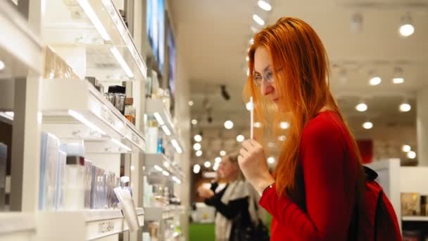 Jovem mulher atraente com cabelo vermelho escolhendo um perfume na área duty free no aeroporto — Vídeo de Stock