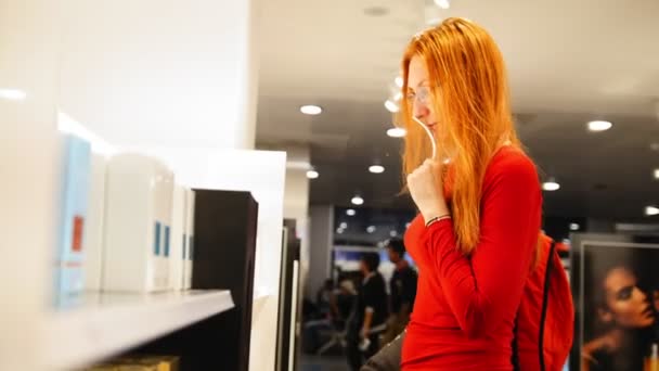 Рыжеволосая девушка в очках выбирает парфюм в зоне беспошлинной торговли аэропорта — стоковое видео