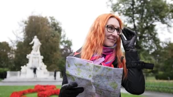 लाल बालों वाली योंग महिला पर्यटक अपने चश्मे को समायोजित करती है और बर्गार्टन, वियना, ऑस्ट्रिया में नक्शे की तलाश करती है — स्टॉक वीडियो