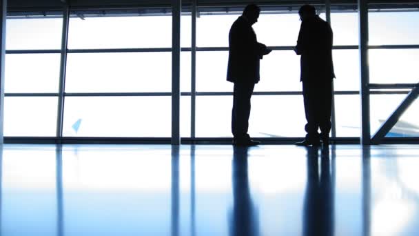 Wo hombres de pie en la terminal del aeropuerto y utilizar los aparatos en frente de la ventana frente a la pista, silueta — Vídeos de Stock