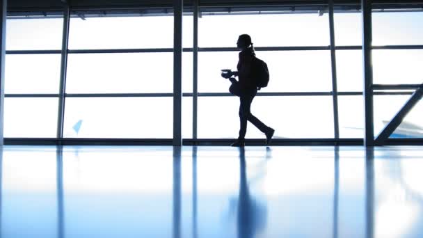 Flickan med ryggsäcken med glasögon kommer i flygplats framför fönstret mittemot landningsbanan, siluett — Stockvideo