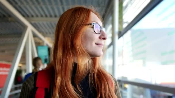 Aeropuerto - mujer feliz con el pelo rojo y gafas tomando la escalera mecánica y mirando a la ventana — Vídeos de Stock