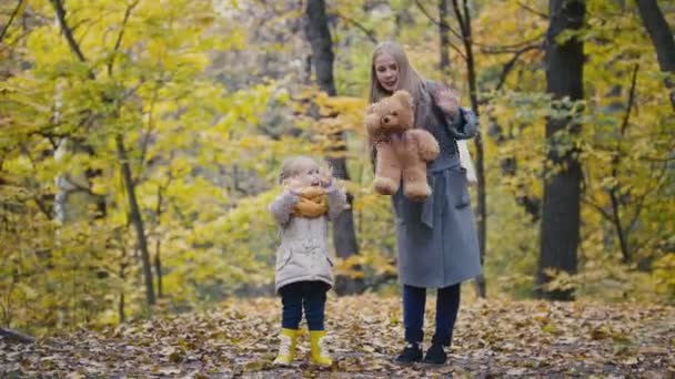 Маленькая дочь с матерью и медвежонком Тедди гуляющими в осеннем парке - машут руками перед камерой — стоковое видео