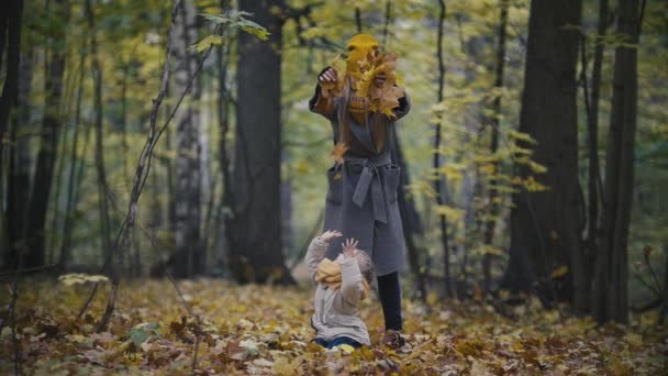 Conceito de família feliz - pequena filha com sua mãe brinca com folhas amarelas no parque de outono, câmera lenta — Vídeo de Stock