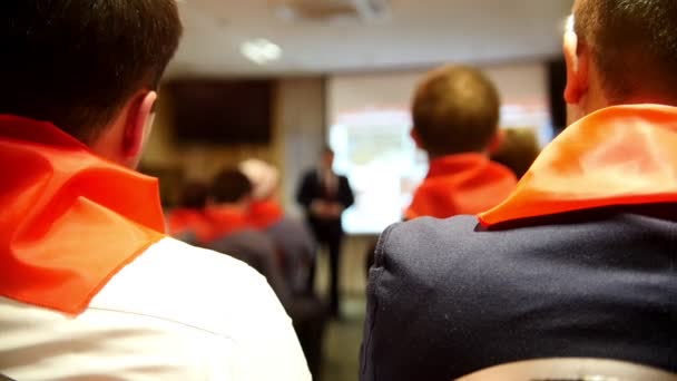 Лектор рассказывает и показывает презентацию на экране для слушателей в красных галстуках — стоковое видео