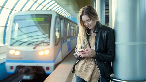 Attraktiva flicka med gadget långa blonda hår i skinnjacka rätar stående i tunnelbanan mot bakgrund av ett tåg som kommer — Stockfoto