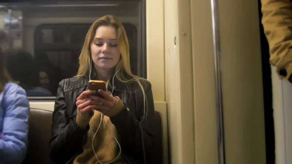 Atractive happy girl with long blonde hair in leather jacket  straightens  use gadget in metro