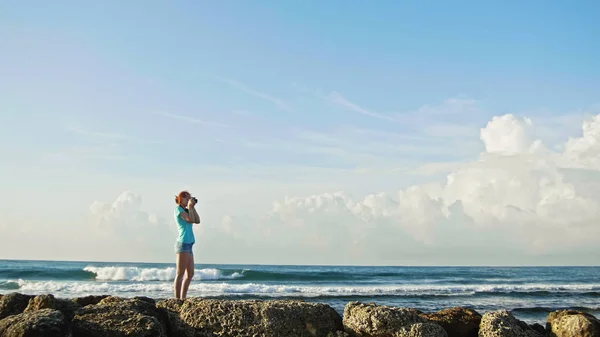 Jovem mulher com cabelo vermelho em óculos fotografias mar em pé sobre as rochas, praia da República Dominicana, grande ângulo — Fotografia de Stock