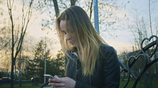 Carino giovane ragazza utilizza il telefono seduto sulla panchina al tramonto — Foto Stock