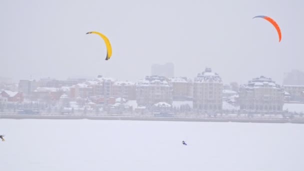 Cerfs-volants de neige jaune et orange sur la rivière de glace en face de la ville moderne - sports extrêmes d'hiver au blizzard — Video