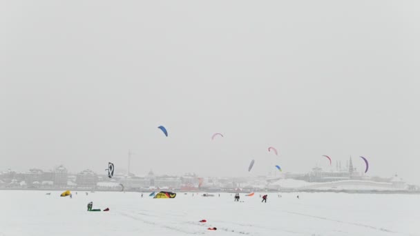 Extremsport im Winter - viele Snowkitesurfer fahren bei bewölktem Himmel auf dem Eisfluss vor der Stadt — Stockvideo