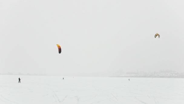 Winter extremal sport - snow-kites over the ice river at snowfall cloudy day — Stock Video