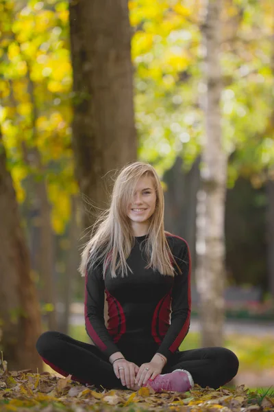 Молодая привлекательная спортсменка, сидящая скрестив ноги на тренировке в осеннем парке, улыбаясь и позируя — стоковое фото