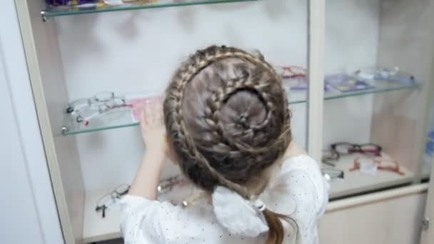 बच्चों के चश्मे ऑप्थाल्मोलॉजी क्लिनिक में छोटी सुनहरे बालों वाली लड़की चश्मा चुनती है — स्टॉक वीडियो