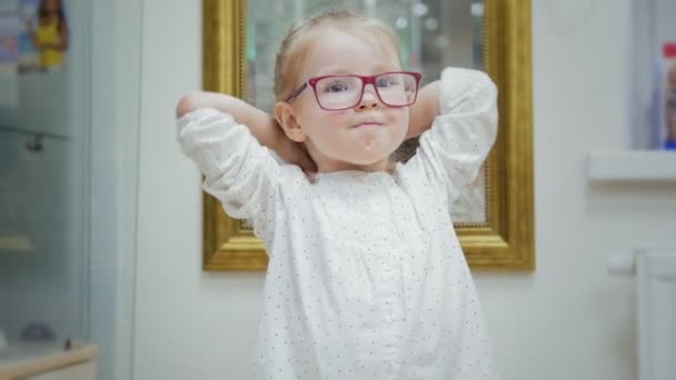 Маленькая девочка пробует очки возле зеркала - шоппинг в офтальмологической клинике — стоковое видео