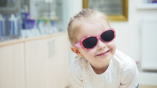 Маленькая девочка играет и Хэмминг перед зеркалом и пытается модные медицинские очки возле зеркала - покупки в офтальмологической клинике — стоковое видео