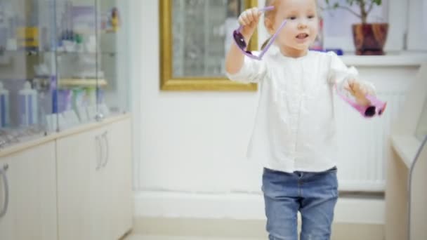 Ребенок - блондинка танцует и просит маму купить красочные очки в медицинском магазине - покупки в клинике — стоковое видео