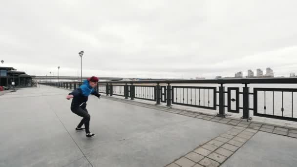Акробатический подросток, выполняющий акробатические сальто и кувыркания на набережной - кепка вылетает из его головы - стедикам замедленной съемки — стоковое видео
