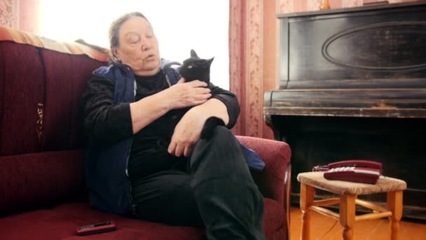 Retrato de senhora sênior em casa - mulher velha senta-se no sofá com gato preto — Vídeo de Stock