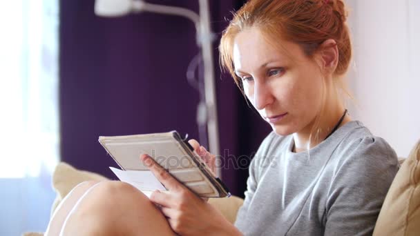 Retrato de una mujer bonita en casa sentada en el sofá y trabajando con gadget y notebook — Vídeos de Stock